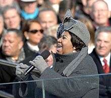 A picture of an African-American woman singing into a microphone that she is holding with her left hand. She is wearing a light grey hat and gloves with a dark grey coat. People can be seen sitting in the background.