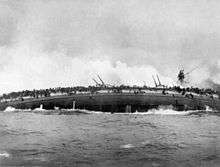 A sinking gray warship rolls over onto is side, its guns pointed skyward