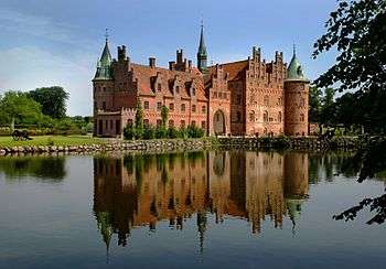 A large three-storied castle has steeply pitched roofs, two circular towers and a spire, and a large arched entrance facing a lake which reflects the castle's image.