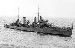 A two funnelled cruiser at sea. People are clustered on the forward deck