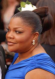 An African American female with dark brown hair up in a bun with a flower. She is wearing a short sleeved royal blue dress.