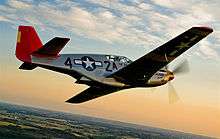 A single nosed-propeller aircraft is in mid-flight above land.  The red nose faces right and the underside of the aircraft is slightly in view.  The aircraft has black propellers, gray base paint, and black letters reading: "A42".  The "4" and "2" are separated by a United States roundel in black with a central white star.  The roundel is also visible on the tops of the wings.  The full length of the right side of the aircraft is visible.