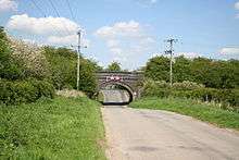 Blue brick single arch bridge carrying former railway over the road into the village. The first houses are just visible through the arch. The road surface has been lowered to pass under the bridge. On either side steep embankments carry the former line, completely overgrown with trees and shrubs.