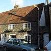 A small, two-storey flint cottage with a steep tiled roof, attached to another of the same height to the left. It has a white door and four irregularly spaced windows (edged in red brick), the upper two of which have window-boxes. A white gate is set into a flint wall attached to the right side of the cottage.